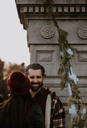 A smiling bearded man in a plaid shirt stands near a stone monument decorated with greenery.