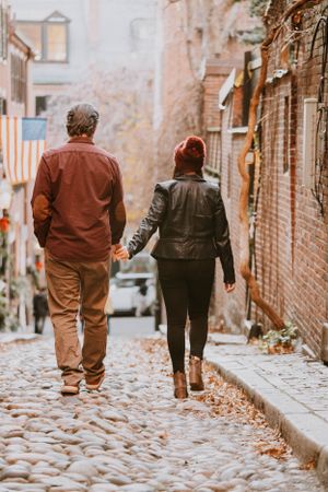 A couple walks hand in hand down a charming cobblestone street lined with brick buildings in autumn.