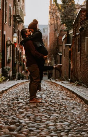 Romantic couple kissing on historic cobblestone street, man lifting woman in red hat.