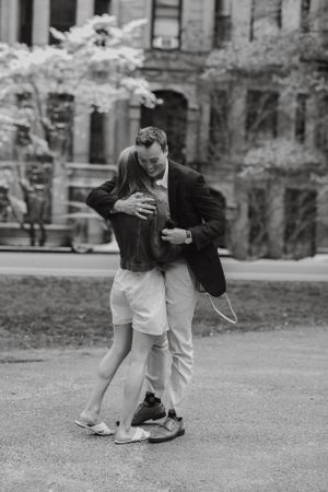 Black and white photo of a couple embracing and dancing outdoors near a blooming tree and historic building.