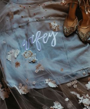 Custom denim jacket with wifey script lettering and floral patches displayed with bridal veil and gold heels.