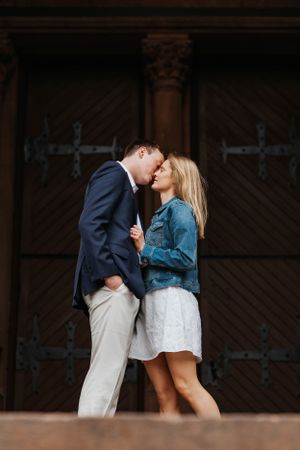 Romantic couple sharing a kiss in front of large decorative wooden doors, woman in denim jacket and white dress.