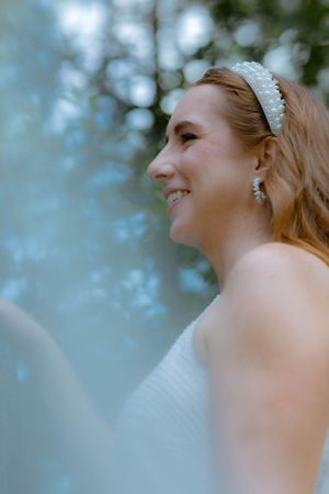 Bride in white dress with pearl headband smiling softly in dreamy outdoor portrait.