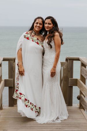 Two women posing on a wooden pier by the ocean, one in a wedding gown and one in a floral white dress.