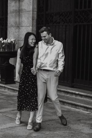 Black and white photo of happy couple walking hand in hand and smiling outside a historic building.