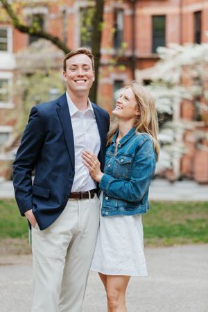 Smiling engaged couple standing together in a blooming spring park near a brick building.