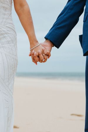 Bride and groom holding hands on a sandy beach, bride wearing a white gown and groom in a blue suit.