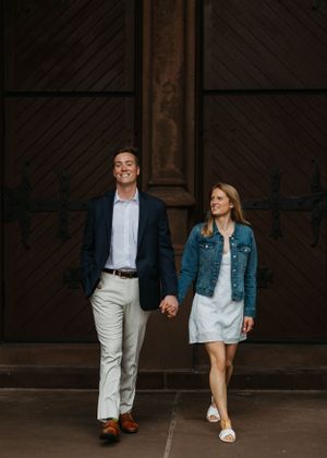 Smiling couple walking hand in hand in front of ornate wooden doors, man in navy blazer and woman in denim jacket.