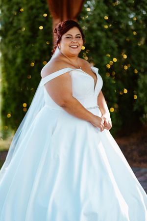 Plus-size bride smiling radiantly in off-shoulder white satin ball gown with crystal belt amid bokeh lights.