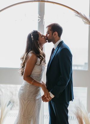 Bride and groom sharing their first kiss at the altar, holding hands in front of a large arched window.