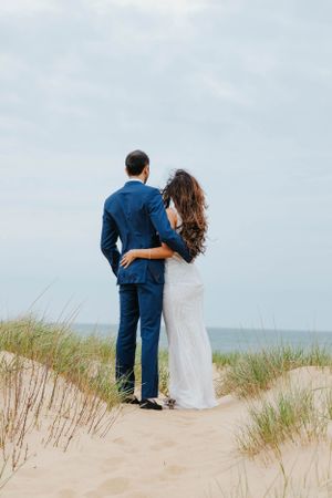 Bride and groom embracing while standing on sandy dunes overlooking the ocean on their wedding day.
