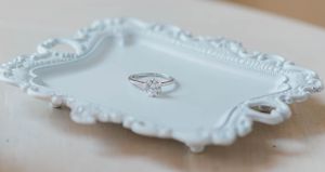 Elegant solitaire diamond engagement ring displayed on a decorative white ornate tray.