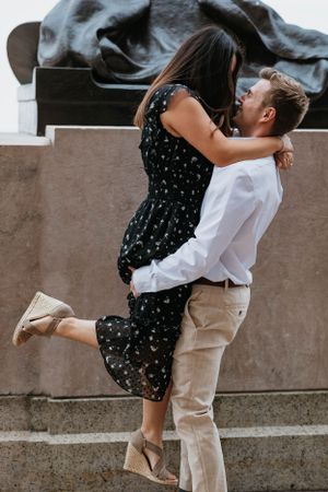 Joyful couple embracing on stone steps, man lifting woman in a black floral dress during engagement photos.