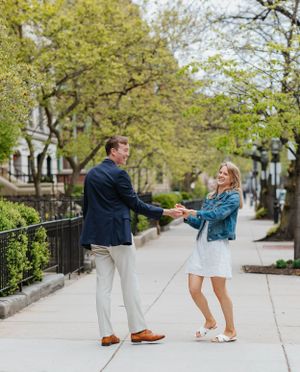 Laughing couple holding hands and swinging on tree-lined city sidewalk, dressed in smart casual spring outfits.