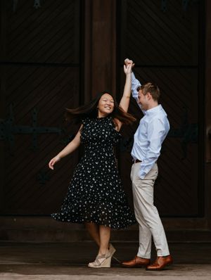 Couple dancing and spinning outdoors near decorative wooden doors, woman in black floral dress laughing with joy.