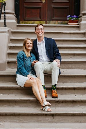 Smiling engaged couple sitting together on brownstone steps, woman showing engagement ring, man in fun printed socks.