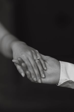 Black and white close-up of couple holding hands showcasing a solitaire diamond engagement ring.