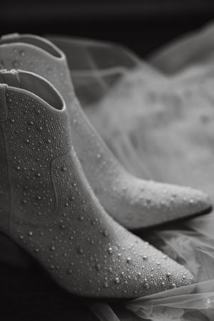 Black and white close-up of pearl-embellished white bridal ankle boots resting on a flowing tulle veil.