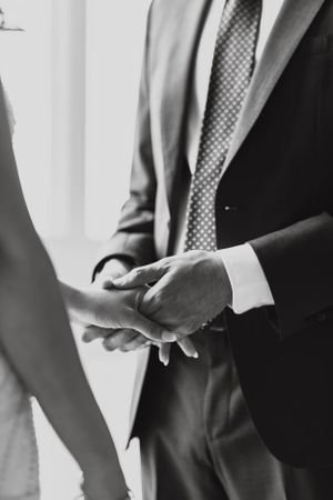 Close-up black and white photo of bride and groom holding hands, groom wearing suit with dotted tie.