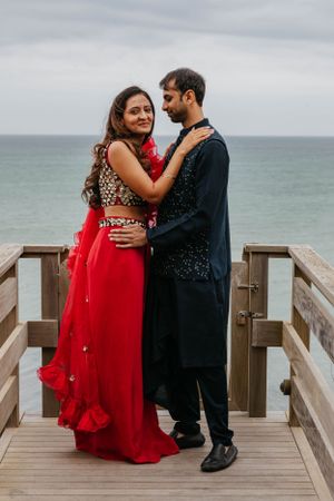 Couple in traditional Indian wedding attire embrace on a wooden boardwalk overlooking the ocean.
