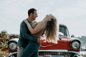 Man dipping and dancing with a woman in front of a classic red vintage car outdoors.