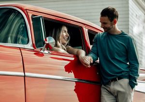 Woman leaning out of red classic car window smiling at man standing beside it.