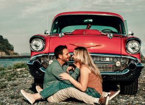 Couple sitting together in front of red vintage Chevrolet car by the water.