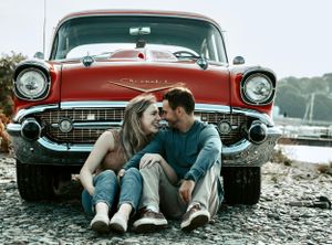 Couple sitting close together leaning against red classic Chevrolet car on rocky shore.