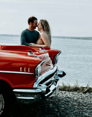 Couple sharing romantic moment leaning over hood of red vintage Chevrolet by the sea.