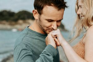 Man gently kissing woman's hand during outdoor engagement photo session by the beach.