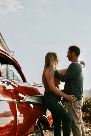 Couple embracing and smiling beside a red classic car in warm sunlight.