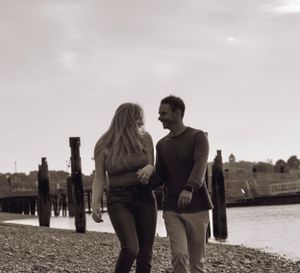 Couple walking hand in hand along a rocky waterfront, sepia-toned engagement photo.