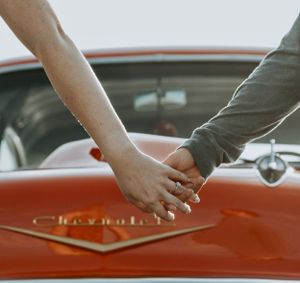 Couple holding hands over the hood of a red vintage Chevrolet car.