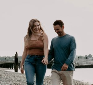 Couple holding hands and laughing while walking along a rocky beach near a pier.