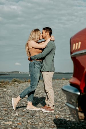 Couple sharing a kiss while embracing near a classic red car on a rocky shoreline.