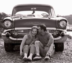 Laughing couple sitting in front of a vintage Chevrolet car in black and white photo.