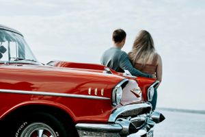 Couple leaning against a classic red vintage car overlooking a calm body of water.
