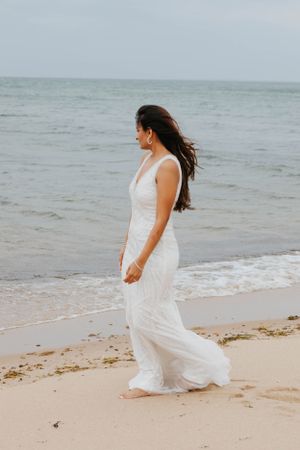 Bride in flowing white beaded gown walks barefoot along a sandy beach with ocean waves behind her.