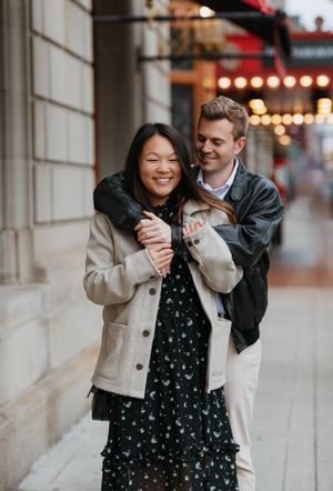 Smiling couple walking together on a city sidewalk, man embracing woman from behind near lit marquee.