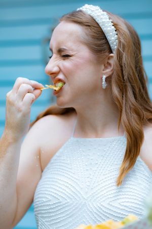 Smiling bride in white pearl dress eating food wearing pearl headband and earrings.