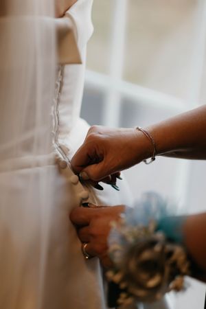 Close-up of hands carefully fastening buttons on back of white satin wedding gown before ceremony.