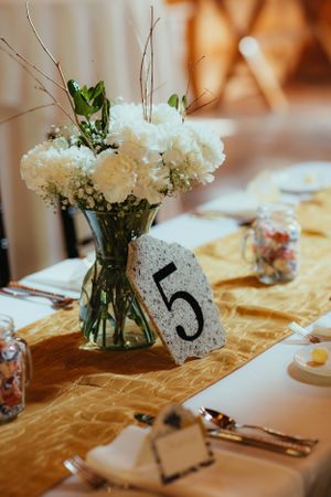 Wedding table number five displayed beside white floral centerpiece on a gold satin table runner.