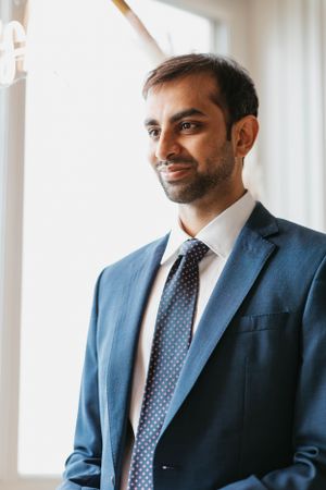 Handsome groom in a blue suit and polka dot tie smiling near a bright window on his wedding day.