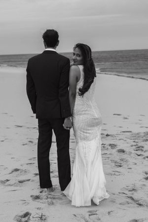 Black and white photo of bride and groom walking on a beach, bride in a fitted white gown looking back.