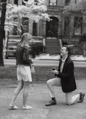 Black and white photo of man on one knee proposing to surprised woman in urban park setting.