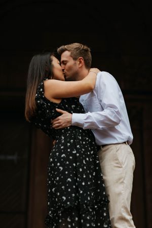 Couple sharing a romantic kiss during their engagement photo session, woman in black floral dress embracing man in white shirt.