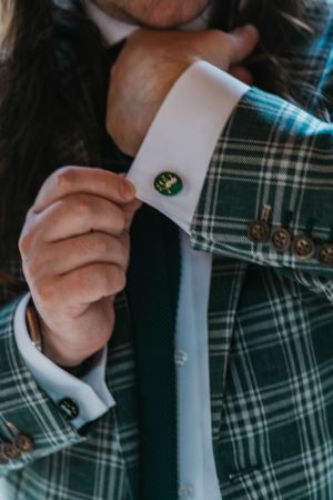 Man adjusting green oval cufflink on white shirt sleeve wearing plaid blazer.