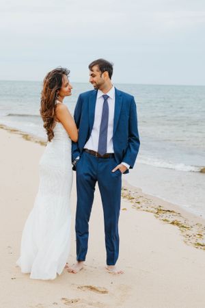 Smiling bride and groom standing barefoot on a beach, groom in a blue suit and bride in a white gown.