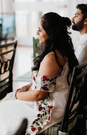 Bride in floral embroidered white dress seated and smiling during wedding ceremony preparations.