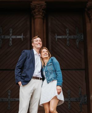 Smiling couple posing together in front of ornate wooden doors, man in navy blazer and woman in denim jacket.